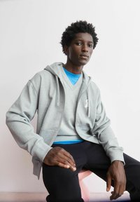 Grijze hoodie met rits en een subtiel logo, gedragen over een blauw shirt en zwarte broek. De stof oogt zacht en gestructureerd.