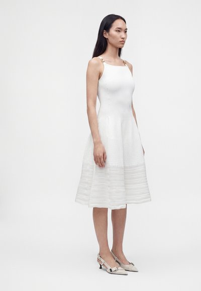 Femme portant une robe midi blanche sans manches avec de fines bretelles et des chaussures pointues beiges à petit talon, debout devant un fond uni.