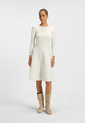 Long-sleeve, cream dress with a fitted bodice and flared hem, featuring a subtle geometric pattern. The dress is paired with beige ankle boots.