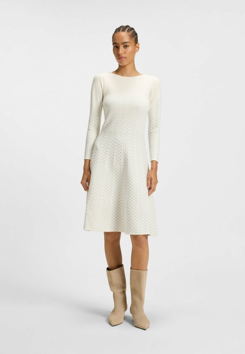 Robe à manches longues, couleur crème, avec un corsage ajusté et un ourlet évasé, affichant un motif géométrique subtil. La robe est associée à des bottines beiges.