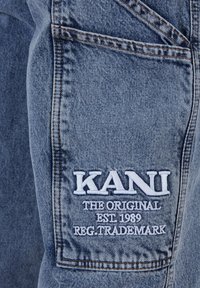 Blue denim fabric shorts featuring stitched details and a prominent white embroidered logo with "KANI THE ORIGINAL EST. 1989 REG. TRADEMARK."