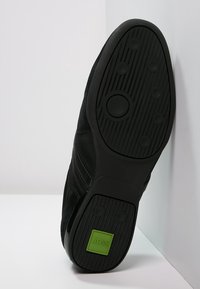 Musta kenkäsole, jossa on pyöreitä pidennyspintoja ja vihreä neliölogo, jossa lukee "BOSS HUGO BOSS" kantapään lähellä.