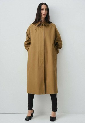 Manteau brun oversize en matériau lisse, doté de poches latérales, d'un col haut et d'une coupe droite. Porté par-dessus des leggings noirs et des talons.