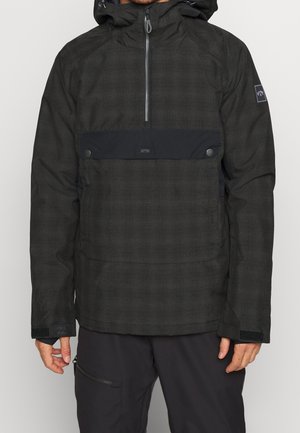 Mężczyzna w czarnej kurtce z kapturem, z przednim zamkiem na pół suwaka, dużą kieszenią na zatrzask oraz paskami na rzepy przy nadgarstkach.