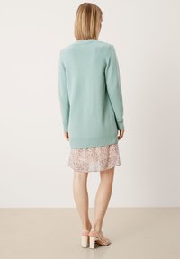 Mátově zelený pletený cardigan s žebrovanými manžetami a lemem, v kombinaci s lehkými šaty s květinovým vzorem. Hladká textura, dlouhé rukávy.