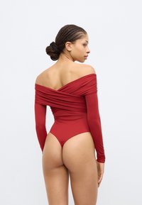 Punane bodi laiade paljastatud õlgadega pikkade varrukatega, mis on volditud detailidega ning kõrge lõikega seljaga. Sile kangas ja vormiv siluett.