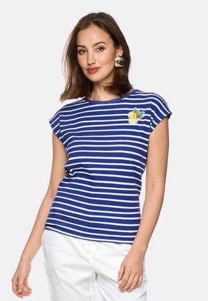 Frau trägt ein blau-weiß gestreiftes T-Shirt mit gestickten Zitronen auf der Brust und weiße Hosen, posiert vor einem schlichten Hintergrund.