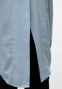 Svijetloplava košulja od glatkog materijala, s bočnim prorezom i zaobljenim donjim rubom, u kombinaciji s crnim trapericama.