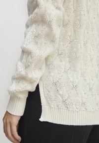 Cremfarbener Strickpullover mit geometrischem Muster, gerippten Bündchen und seitlichen Schlitzen für zusätzliche Details und Textur.