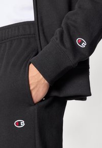 Μαύρο φούτερ και παντελόνι από απαλή ύλη, με ριμπ μανσέτες και πλαϊνές τσέπες. Διακριτικό κεντητό λογότυπο στο μανίκι και το πόδι.