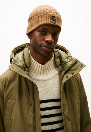 Tan gestrickte Mütze mit Rippenstruktur und Logo, grün gefütterte Kapuzenjacke, cremefarbenem Pullover mit horizontalen schwarzen Streifen.