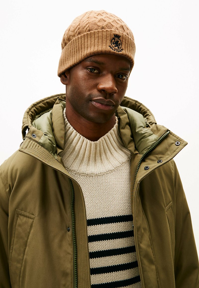 Tan gestrickte Mütze mit Rippenstruktur und Logo, grün gefütterte Kapuzenjacke, cremefarbenem Pullover mit horizontalen schwarzen Streifen.