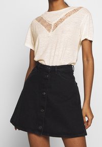 Mujer vistiendo una blusa corta de manga con detalles de encaje en color beige, metida en una falda de mezclilla negra de tiro alto con botones en la parte delantera.