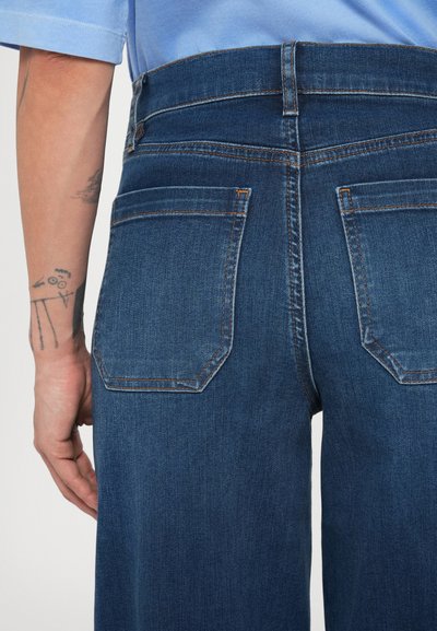 Sötétkék magas derekú farmernadrág, két hátsó zsebbel és kontrasztos varrással. A szövet sima textúrájú.