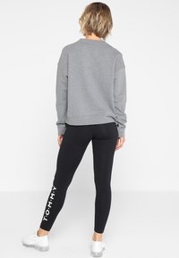 Persoon in een grijze sweatshirt en zwarte leggings met de tekst "TOMMY" op het linkerpijp, staande met de rug naar de camera tegen een witte achtergrond.