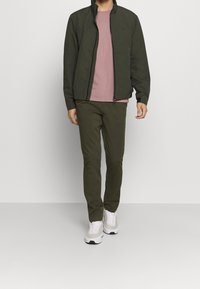 Vihreä vetoketjutakki, vaaleanpunainen paita, oliivinvihreät housut ja vaaleanharmaat lenkkarit. Materiaalit vaikuttavat sileiltä, minimalistinen muotoilu.