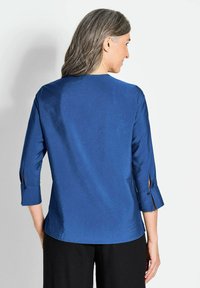 Blusa in raso blu con maniche a tre quarti, caratterizzata da un collo rotondo e dettagli con bottoni ai polsini, texture liscia e leggera lucentezza.