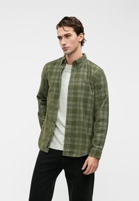 Oliven grønn rutete skjorte med krage, lange ermer og en lomme. Brukt over en lysegrå T-skjorte og svarte jeans.