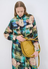 Camicia oversize a fantasia floreale con una palette di colori verde e nero, abbinata a una borsa a tracolla rotonda in pelle gialla con dettaglio a nodo.