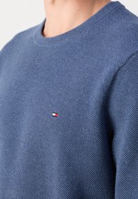 Maglione lavorato a maglia blu con collo a giro, tessuto testurizzato e una piccola etichetta logo rossa, bianca e blu sul lato sinistro del petto.