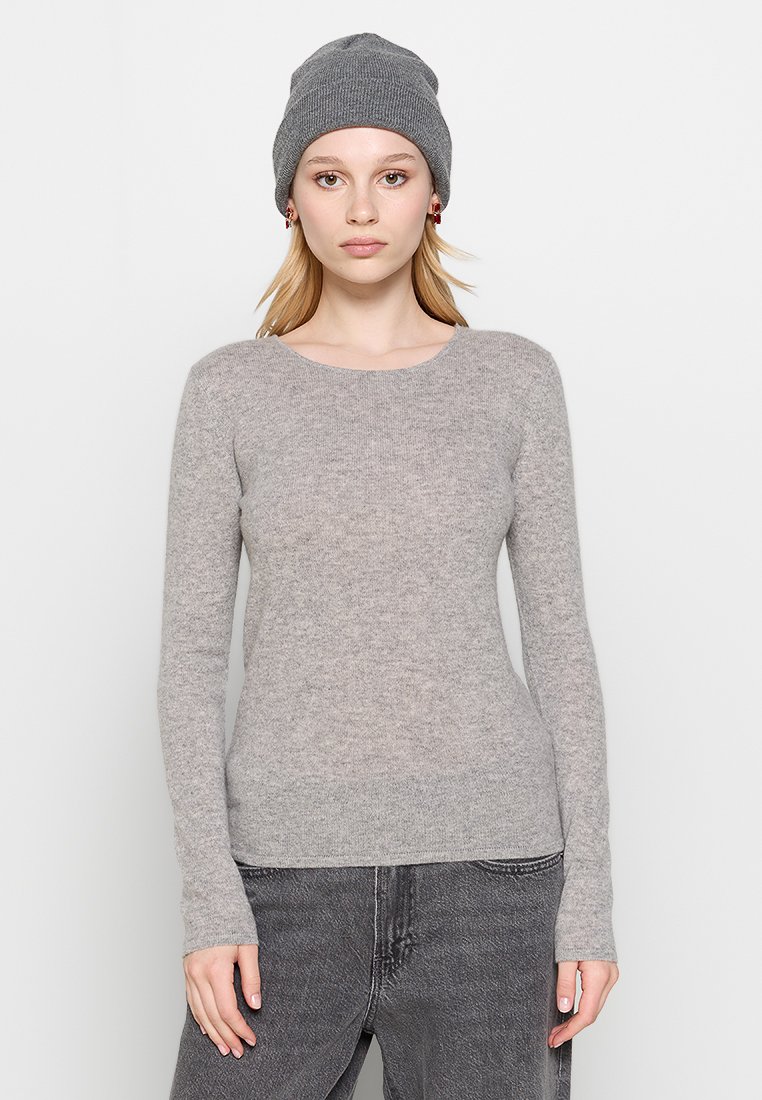 Kobieta o jasnej karnacji i blond włosach, nosząca szary wełniany beret, jasnoszare długie rękawy sweter i ciemnoszare dżinsy, zwrócona przodem z neutralnym wyrazem twarzy.