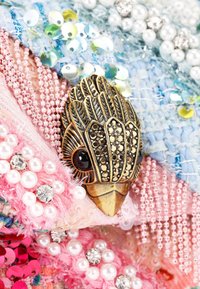 Broche en bronze en forme de tête d'oiseau, ornée de pierres noires et de perles, posée sur un fond texturé de tissu coloré et de paillettes.