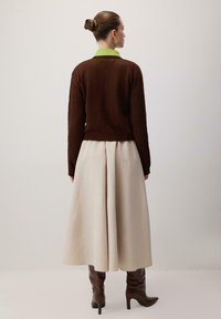 Brauns adīts džemperis, bēša plisētā svārki un brūnas augstpapēžu zābaki. Zaļš apkakle parādās no zem džempera. Gludās tekstūras.