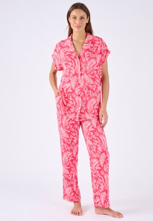 Donna in piedi a piedi nudi che indossa un completo coordinato con camicia a maniche corte e pantaloni, stampati con foglie tropicali rosa e bianche.