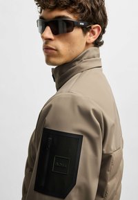 Beige pufferjacka med hög krage, har en svart dragkedjeficka och en svart tygpatch med logotyp på ärmen. Svarta solglasögon.