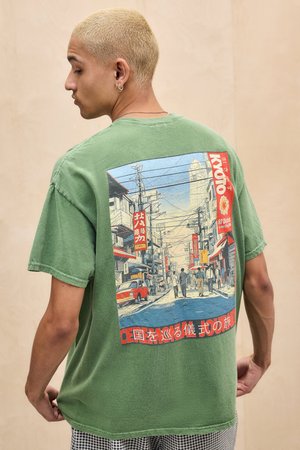 Jeune homme aux cheveux blonds courts portant un t-shirt vert orné d'une scène de rue illustrée avec des panneaux rouges et des gens marchant.