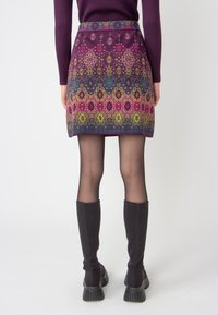 Short patterned skirt in purple, featuring ornate designs in green, pink, and blue, with a straight cut and waistband. Worn with knee-high black socks and chunky shoes.