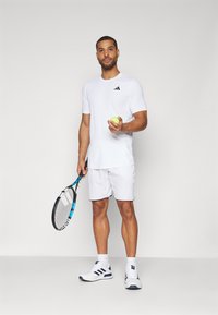 Maglietta sportiva bianca con logo Adidas nero, pantaloni corti bianchi abbinati, tiene una pallina da tennis gialla e una racchetta da tennis con accenti blu.