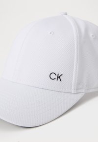 Fehér sapka textúrált anyagból, ívelt karimával és fekete "CK" logóval az elején. Klasszikus kialakítás szellőzőnyílásokkal.
