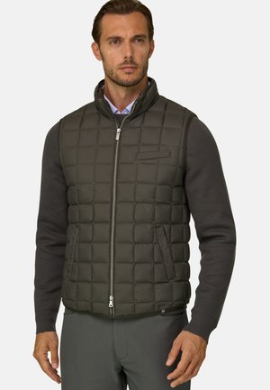 Mann, der eine dunkel olivgrüne gesteppte Weste über einem grauen Langarmpullover und grauen Hosen trägt, mit neutralem Gesichtsausdruck.