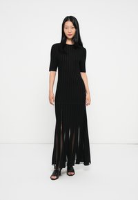 Crna rebrasta maksi haljina s kratkim rukavima, koja ima prozirni donji dio i vertikalne pruge, dopunjena crnim kliznim cipelicama.