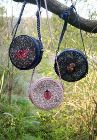 Kolm ümarat risti üle keha kantavat kotti, millel on tikitud putukate ja loomade mustrid, mis ripuvad puuoksa küljes õues.