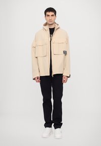 Beige zip-up jacket with two chest pockets and a patch detail, paired with black pants and white sneakers. Neutral tone and casual design.