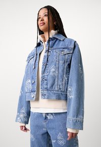 Veste en denim bleu clair coupée avec des motifs graphiques blancs, un col classique, une fermeture à boutons et de grandes poches avant.