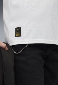 Maglietta da football bianca con un motivo texture, con un'etichetta rettangolare nera con stampa dorata, indossata con abbigliamento scuro e una catena.