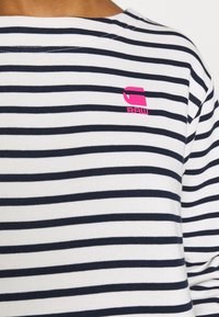 Chemise à manches longues rayée bleu marine et blanche en coton, avec un petit logo rose dans le coin supérieur gauche. Les rayures horizontales sont homogènes.