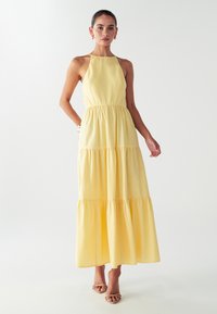 Nainen seisoo hihattomassa keltaisessa kerroksellisessa maksimekossa, kullanväriset korvakorut, rannekoru ja vaaleanbeiget korkokengät, valkoista taustaa vasten.