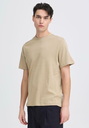 Jonge man met donker haar die een effen beige T-shirt met korte mouwen en een donkere broek draagt, staand tegen een lichte achtergrond.