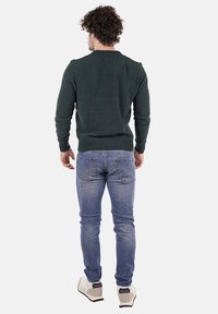 Maglione lavorato a maglia verde scuro con polsini e fondo a coste, abbinato a jeans blu chiaro e sneakers bianche, visto da dietro.