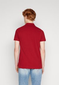 Červené polo tričko s krátkým rukávem a límečkem, s hladkou bavlněnou texturou, přímým střihem a bočními rozparky, nošené s světle modrými džínami.
