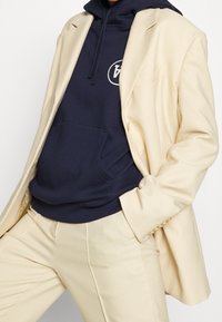 Marinsträv hoodie med ficka framtill, lagd under en krämfärgad blazer, ihop med matchande krämfärgade skräddarsydda byxor. Slät tyg, strukturerad design.