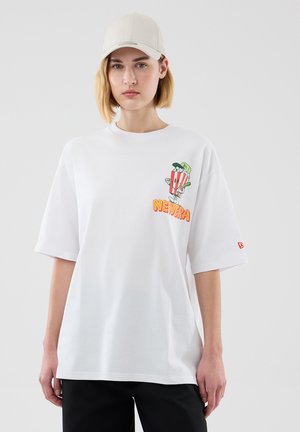 Persona que lleva una camiseta blanca oversize con un personaje de palomitas y el texto "NEW ERA" en el pecho, combinada con una gorra de béisbol beige y pantalones negros.