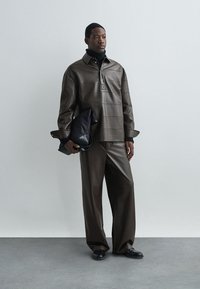 Brun skinnjacka med hög krage och horisontella paneler, kombinerad med matchande lösa byxor. Svart kuvertväska i handen.