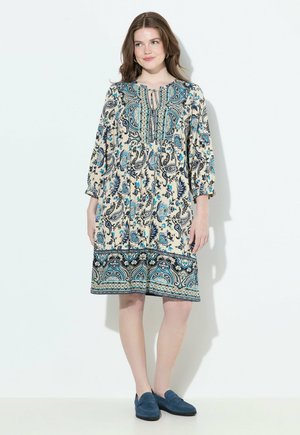 Donna vestita con un abito beige al ginocchio con motivi floreali e paisley blu, maniche a tre quarti e scarpe blu senza lacci, in piedi contro un muro bianco.