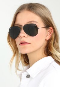 Ochelari de soare aviator negri cu lentile mari negre, cadru metalic și brațe subțiri negre, purtați de un model cu păr blond și o bluză cu guler alb.