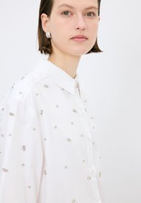 Bílá košile s roztroušenými stříbrnými ozdobami, zapínání na knoflíky a klasický límeček. Hladká textura, pohodlný střih, moderní estetika.